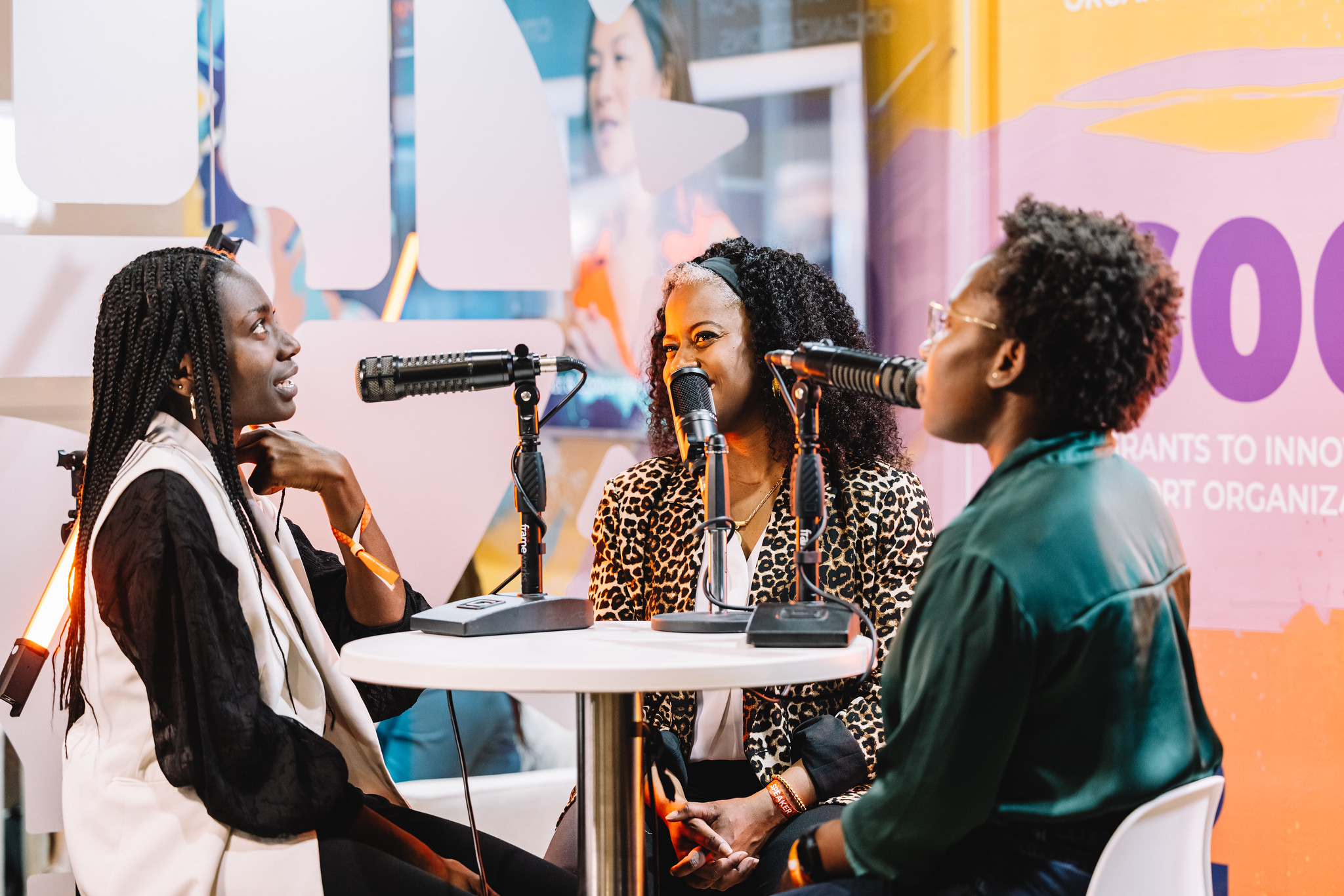 Three people sit at a high table. One appears to be talking, while the other two listen. All three have individual microphones. They appear to be recording a podcast.