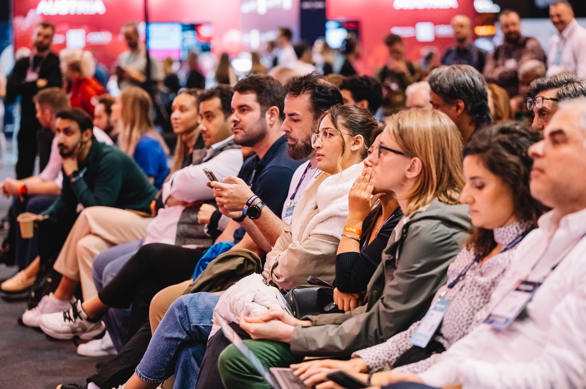 A seated audience of people viewed side on. Most of these people are looking away from camera in the same direction. One person in the centre of the front row of people is looking at their mobile phone, and another person is using a laptop.