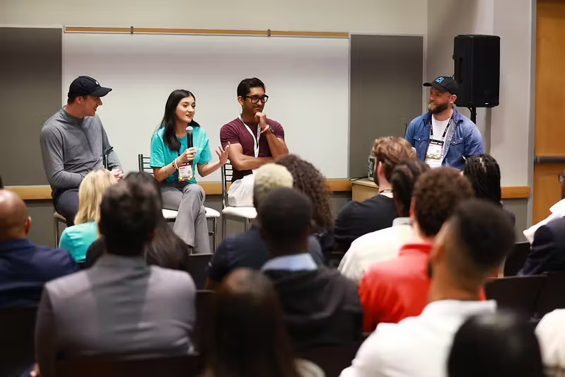 Photograph of a packed room of people. There are three people sitting on chairs on stage. The person sitting in the middle is holding a microphone. On the right side of the stage, there is another person sitting on a chair looking toward the other three people.