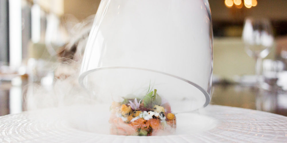 a glass dome lifted from a delicate dish with salmon and herbs, smoke is disipating