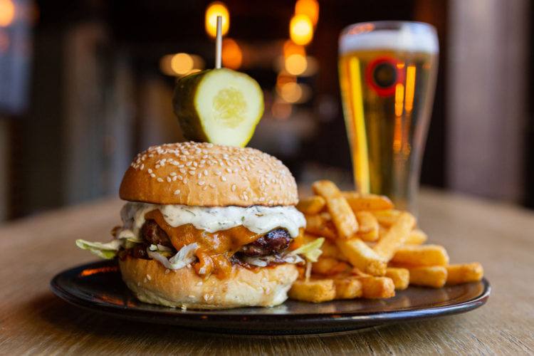 closeup of a tall burger with chunky fries on the side and a pint of lager in the back