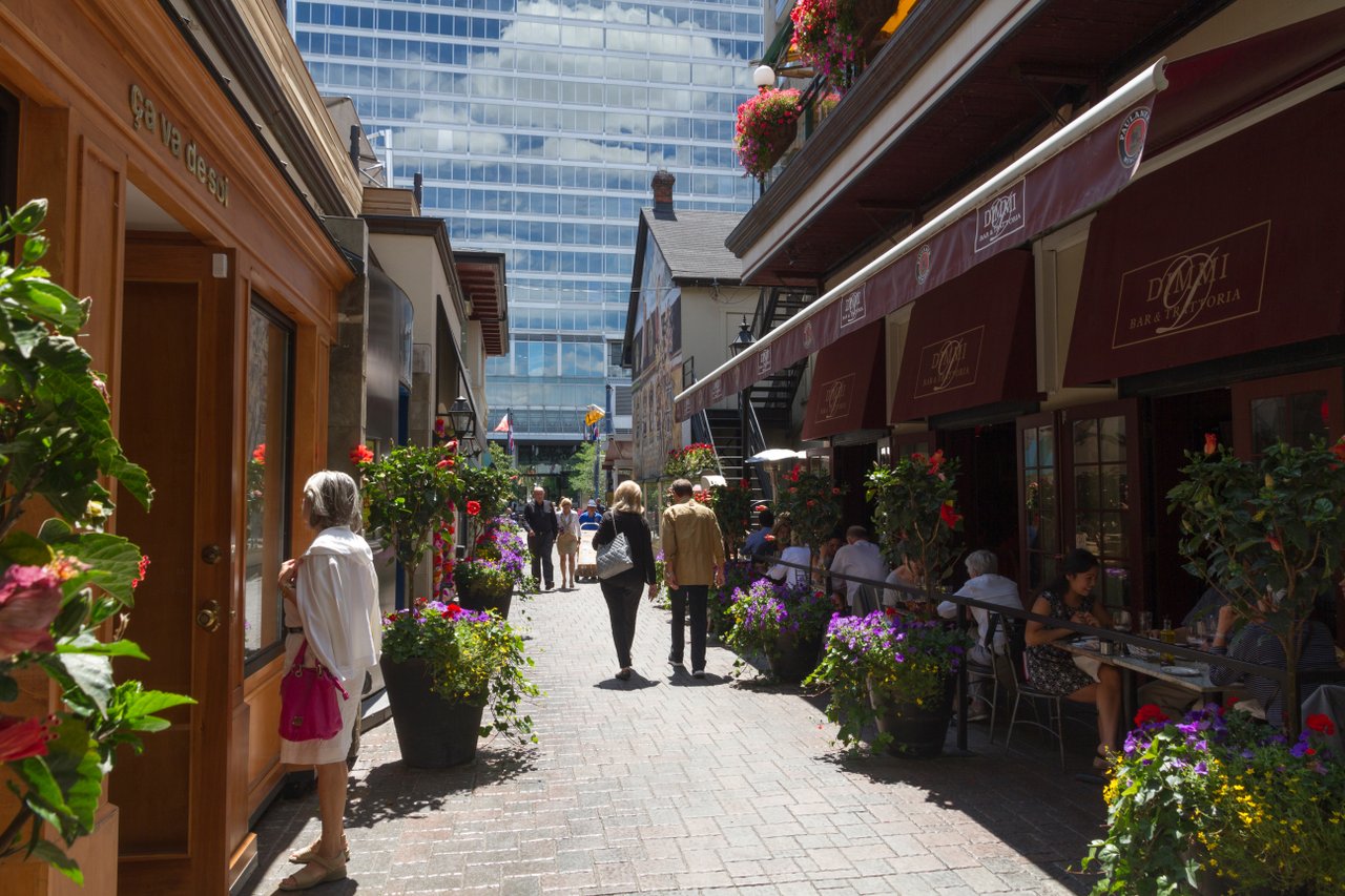 Corridor of shops on a sunny day. People smiling and strolling. There are many flower pots sprinkled around the shop fronts.